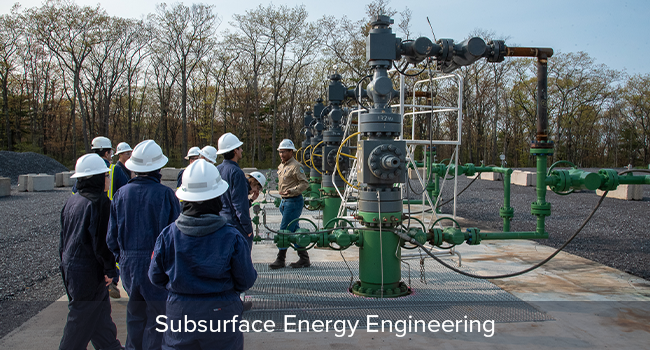 Student group wearing hardhats at energy facility looking at industrial pipes.