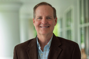 Russell Johns in blue shirt and brown suit smiling.